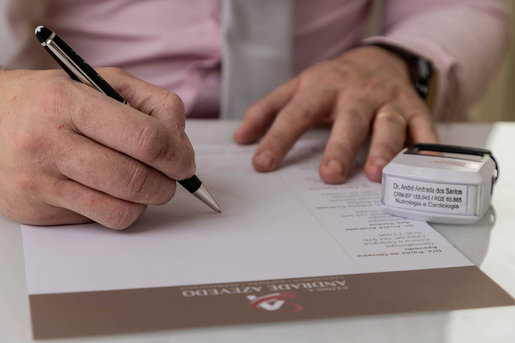 Mãos do Dr. André escrevendo uma prescrição em papel timbrado da clínica